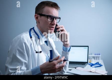 European doctor in white medical coat and glasses consults patient on cell phone in clinic office, sitting at table with laptop. German general Stock Photo