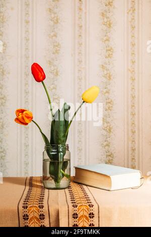Three red and yellow tulips in the glass jar and a book. Retro vibes. Copy space Stock Photo