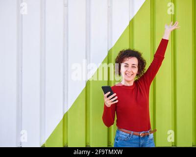 A curly-haired woman who wears a sweater and jeans, raises one hand while in the other she has a mobile phone, in the background there is a green and Stock Photo