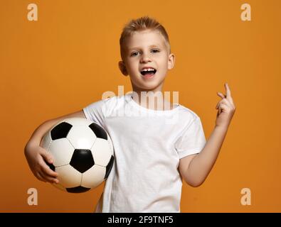 Active sporty blonde kid boy in white blank t-shirt stands holding soccer ball in hand and gestures one, first sign Stock Photo