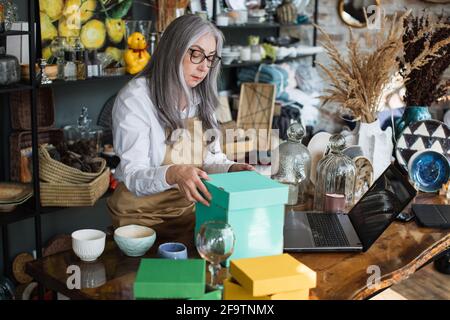 Attractive aged woman in eyeglasses and apron doing inventory at decor store. Competent saleswoman rechecking all available assortment at shop. Stock Photo
