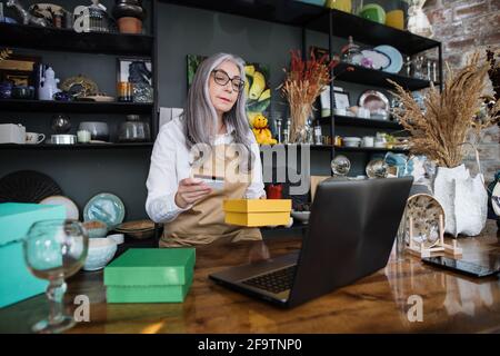 Professional aged woman holding discount card and yellow box while sitting at decor store. Saleswoman using modern laptop at workplace. Stock Photo