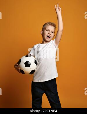 Screaming loudly blonde kid boy in white blank t-shirt stands holding soccer ball in hand and gestures V victory sign Stock Photo
