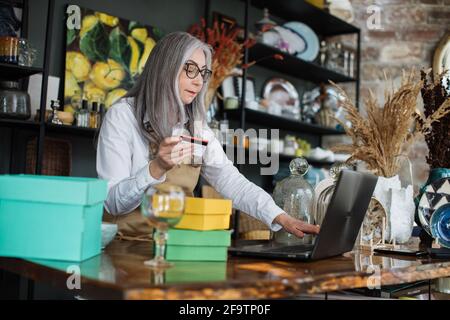 Shopping and discount system concept. Likable grey-haired lady typing on modern laptop while sitting at counter with credit discount card in hand. Shelves with various beautiful decor on background. Stock Photo