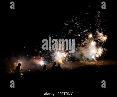 Fireworks explode all over beach in nighttime blackness with people in silhouette. Stock Photo