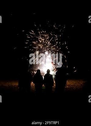 Group of people with back to camera in silhouette have a close up view of fireworks exploding on beach in black night. Stock Photo
