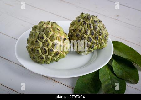 Two sugar apple with leaves on a white plate on white aged wooden table. Stock Photo