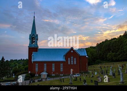 The sun sets on a cloudy evening behind a red church on a grassy hill surrounded by a cemetery Stock Photo