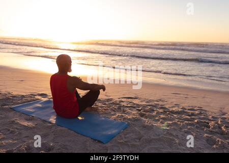 Senior african american man meditating and practicing yoga while sitting on yoga mat at the beach Stock Photo