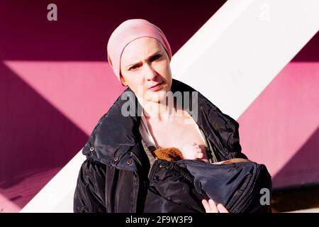 portrait of a woman sick with cancer carrying her baby with a headscarf walking down the street with a pink wall in the background. concept fight agai Stock Photo