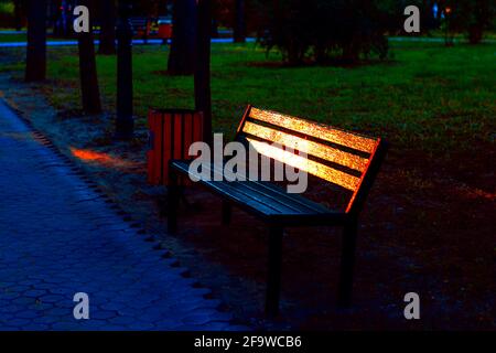 Bench in the evening park with light reflection in the painted planks . No people in the park Stock Photo
