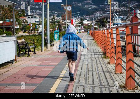 The little boy in jeans jacket traveler walks on a tourist promenade with beautiful shrubs, near the sea, boats and ships on the background of the cit Stock Photo