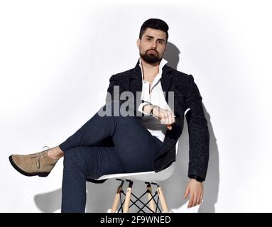Young stylish bearded man in jeans and casual tweed jacket is sits on chair in relaxed pose with his legs crossed Stock Photo