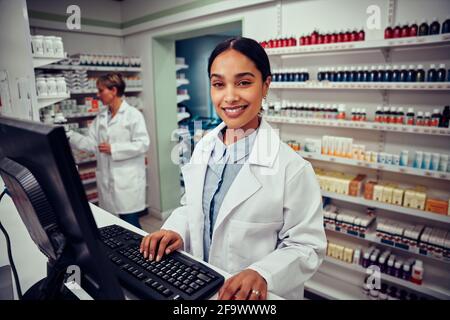 Portrait of smiling young female pharmacist wearing labcoat working on computer in drugstore looking at camera Stock Photo