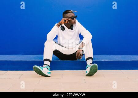 Young afro American black man wearing a white sweatshirt, basketball sneakers, sunglasses, comb in the hair and headphones seated on a blue wall looki Stock Photo