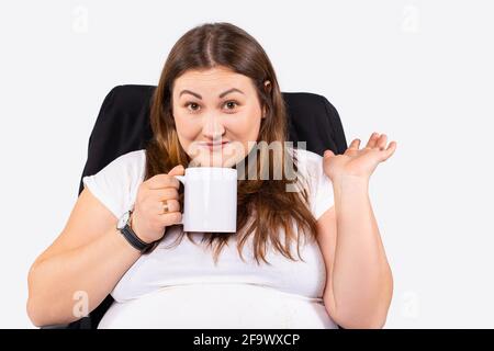 Portrait of a beautiful smiling young overweight blonde large woman wearing white blouse standing isolated over grey background, holding tea cup with Stock Photo