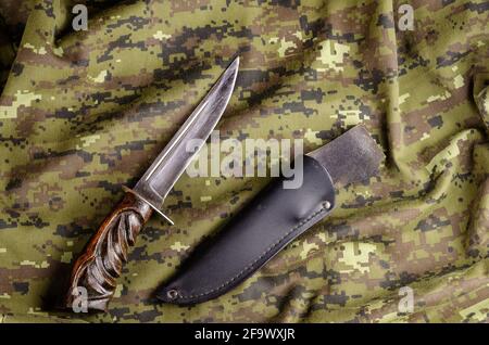 Knife with wooden handle and leather scabbard on camouflage fabric. Extracted from the scabbard is a large knife lying on a military uniform. Army the Stock Photo