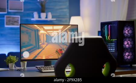 RGB powerful personal computer gamer rig with first-person shooter game on screen. Monitor stands on table in home gaming room. Cozy room with modern design is lit with warm and neon light. Stock Photo
