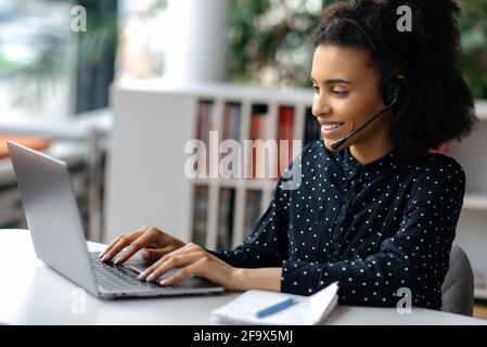 African American beautiful young woman wearing a headset, sitting at a work desk, using a laptop, working online, surfing the Internet, chatting with colleagues or clients, answers to email, smiling Stock Photo