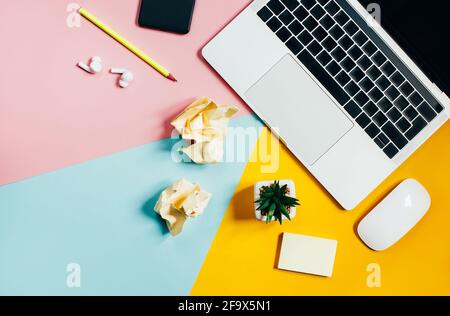 top view of a laptop, computer mouse, mobile phone, pencil, cactus, crumpled pieces of paper on a yellow, pink and blue background with free space for Stock Photo