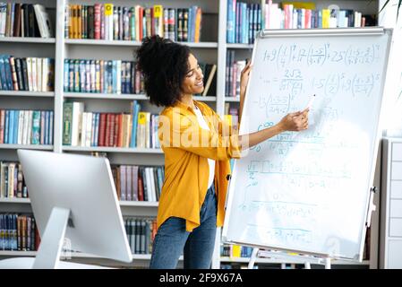 Distance training, online lecture. Young smart african american female teacher conducts online lesson for students via video conference uses computer, stand at library, shows information on whiteboard Stock Photo