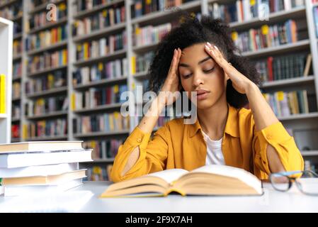 A clever focused african american millennial female student sitting in a college library at table with laptop, reading book with interest, preparing for lecture or exam, knowledge and learning concept Stock Photo