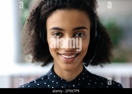 Close-up of African American charming confident young woman in headset, sitting in office, working as operator of call center or support service, looks directly at the camera and smiling friendly Stock Photo