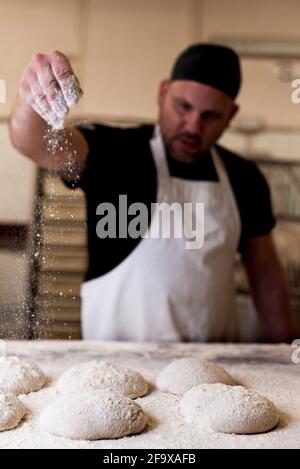young baker in bakery shop sprinkling flour with hands on fresh bread dough in front of oven. concept of traditional manual bread preparation in bread Stock Photo