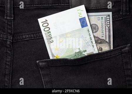 One hundred euro and one hundred dollar bills sticking out of the pocket of black jeans Stock Photo