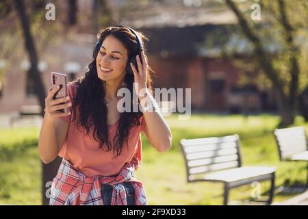 Happy young woman is enjoying sunny day outdoor in city. She is listening music on headphones. Stock Photo