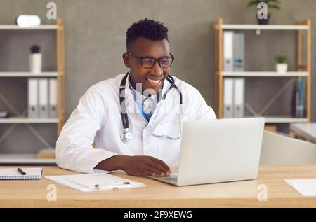 Happy black doctor sitting at office desk and using laptop for video call with patient Stock Photo