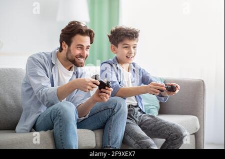 Boys are boys. Joyful father and teen son competing with each other in online video games, using joysticks Stock Photo