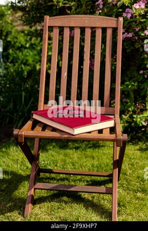 A large hardcover book resting on a chair with a pair of reading glasses placed on top. Reading outdoors concept Stock Photo