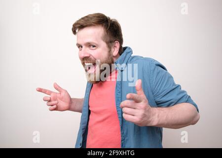 Funny bearded man in casual clothes makes a gesture with hands, smiles broadly and looks slyly at the camera. White background. Stock Photo