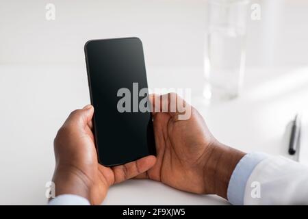 Online doctor and remote advice. Black male doctor using smartphone with blank screen, consulting patients distantly Stock Photo