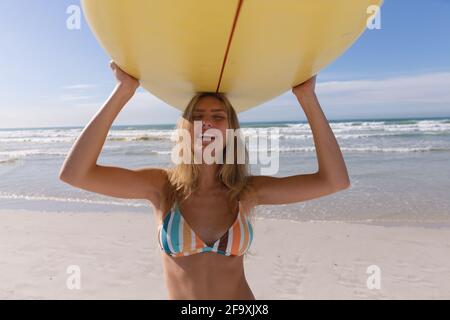 Smiling caucasian woman wearing striped bikini carrying yellow surfboard on her head at the beach Stock Photo