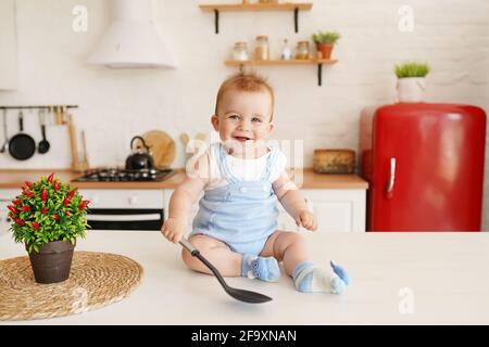 Cute child baby boy 1 years old sitting on the table with big spoon in the kitchen room Stock Photo