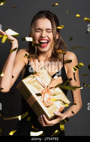 excited young woman in black slip dress holding gift box near blurred confetti on grey Stock Photo