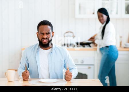 Starving Angry Black Man Waiting For Food At Table In Kitchen Stock Photo