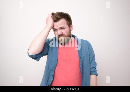 Young bearded man suffers from severe headache or is under stress, he holds forehead with hand and looks at the camera.  Stock Photo