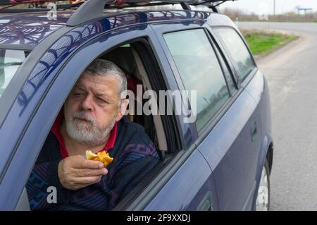 Hungry Caucasian senior driver eating patty with apple inside his car on short stop on roadside while traveling in Ukraine Stock Photo