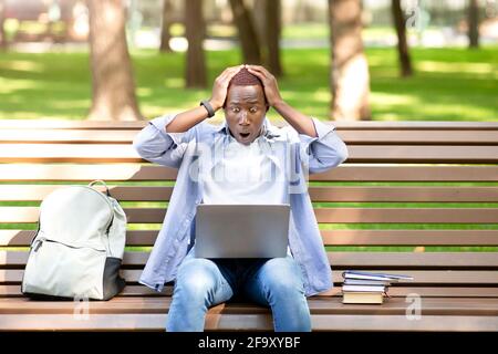 Terrified black man looking at laptop screen and grasping his head in panic on bench at park Stock Photo