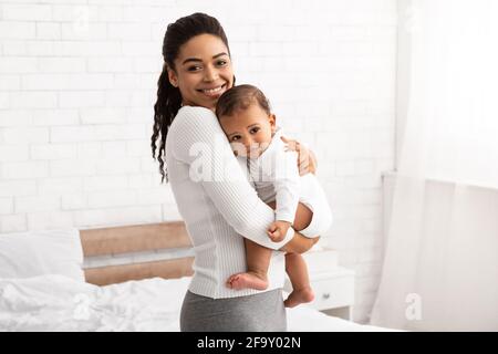 African American Mommy Carrying Little Baby Standing In Bedroom Indoor Stock Photo