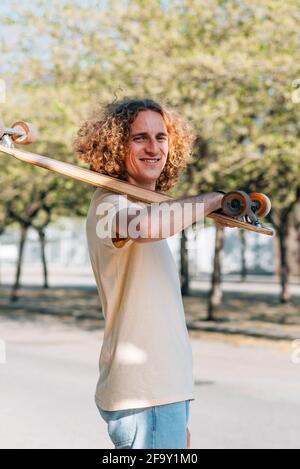 vertical take of a caucasian man holding a skateboard in the street. He has curly long hair and dresses casual clothes. He looks at camera smiling Stock Photo