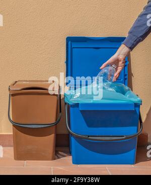 A hand puts an empty plastic bottle into a blue recycling bin, with another brown bucket next to it Stock Photo