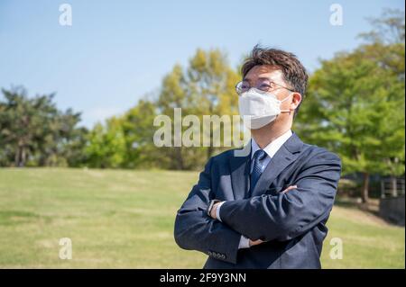 Asian middle-aged businessman with folded arms wearing a mask on the lawn. Stock Photo