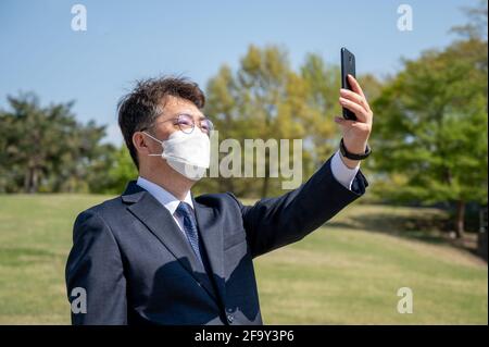 Middle-aged Asian businessman wearing a mask and using a smartphone on the lawn. Stock Photo