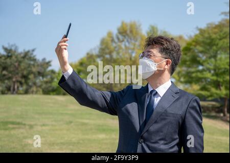 Middle-aged Asian businessman wearing a mask and using a smartphone on the lawn. Stock Photo