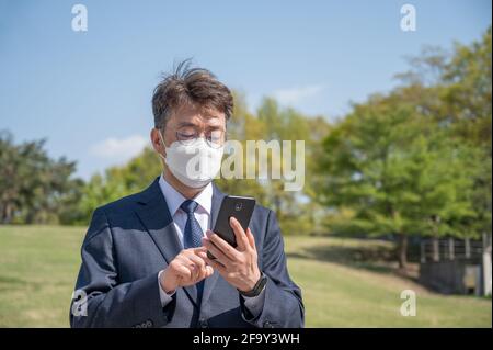 Middle-aged Asian businessman wearing a mask and using a smartphone on the lawn. Stock Photo