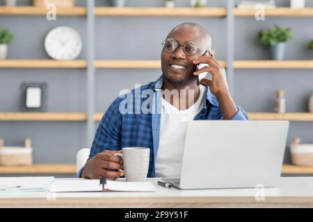 Coffee break. Happy african american employee talking on cellphone and holding cup with hot drink Stock Photo